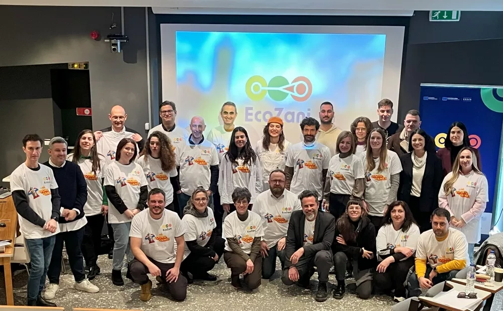 Group photo of the EcoZani project team, including local stakeholders and international partners, standing together indoors in front of a screen with the EcoZani logo. Many participants are wearing white project T-shirts and smiling at the camera.