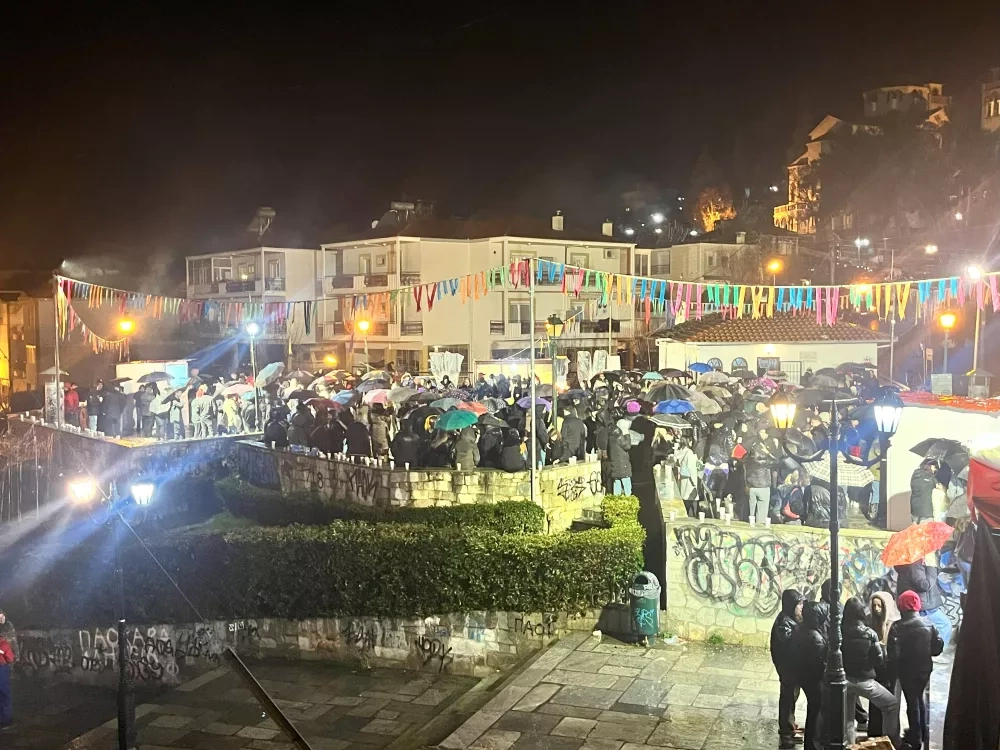 A rainy night festival: crowds huddle under umbrellas on stone terraces, colorful pennant banners overhead, lamplight reflecting off wet pavement in a small town square.