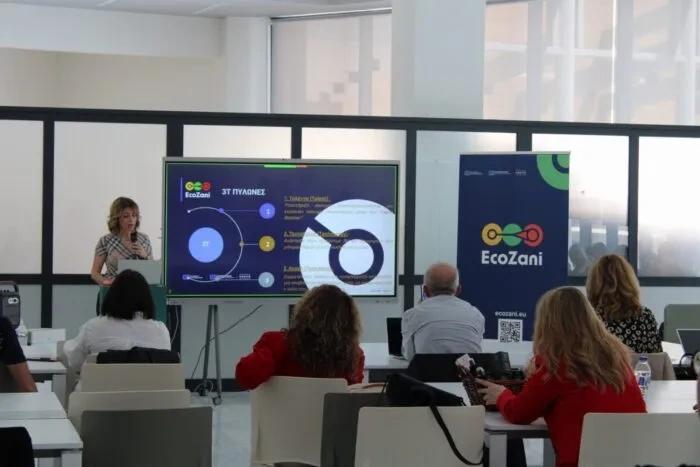 A speaker presents the EcoZani project during a workshop, standing beside a large screen with slides, while participants sit at tables in a modern conference room with EcoZani branding visible.
