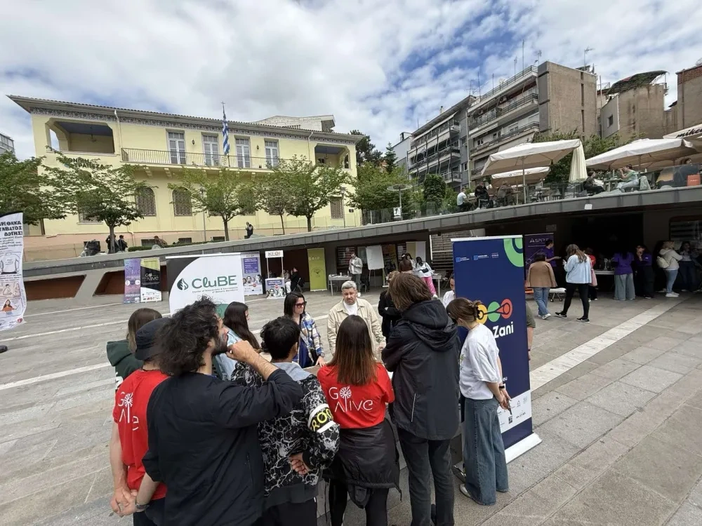 Participants gather in a public square during an outdoor EcoZani event, engaging in discussion around an information stand, with city buildings and project banners visible in the background.