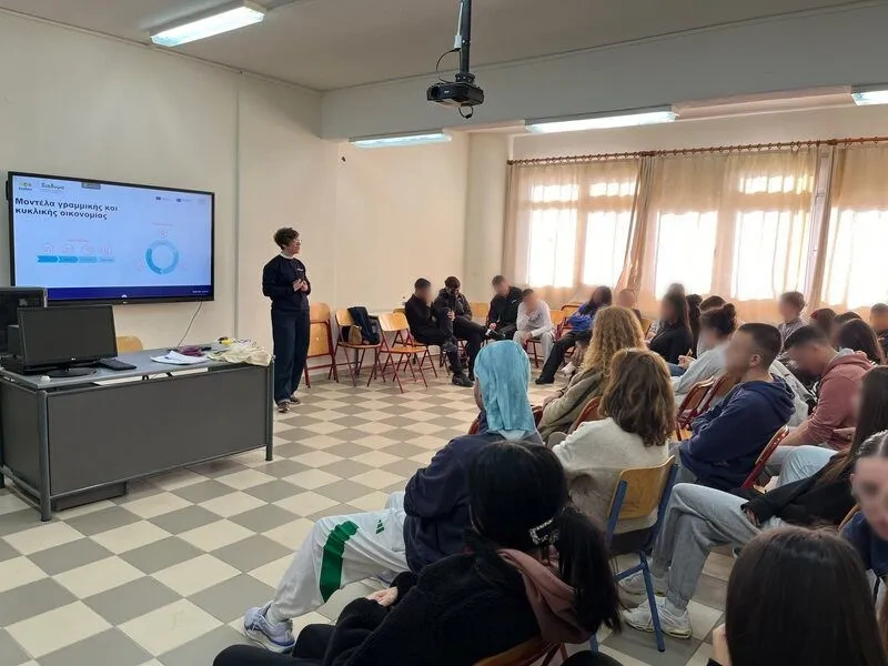 A facilitator delivers a presentation on circular economy concepts to a group of students seated in a classroom, with a screen displaying slides and participants listening attentively during an educational workshop