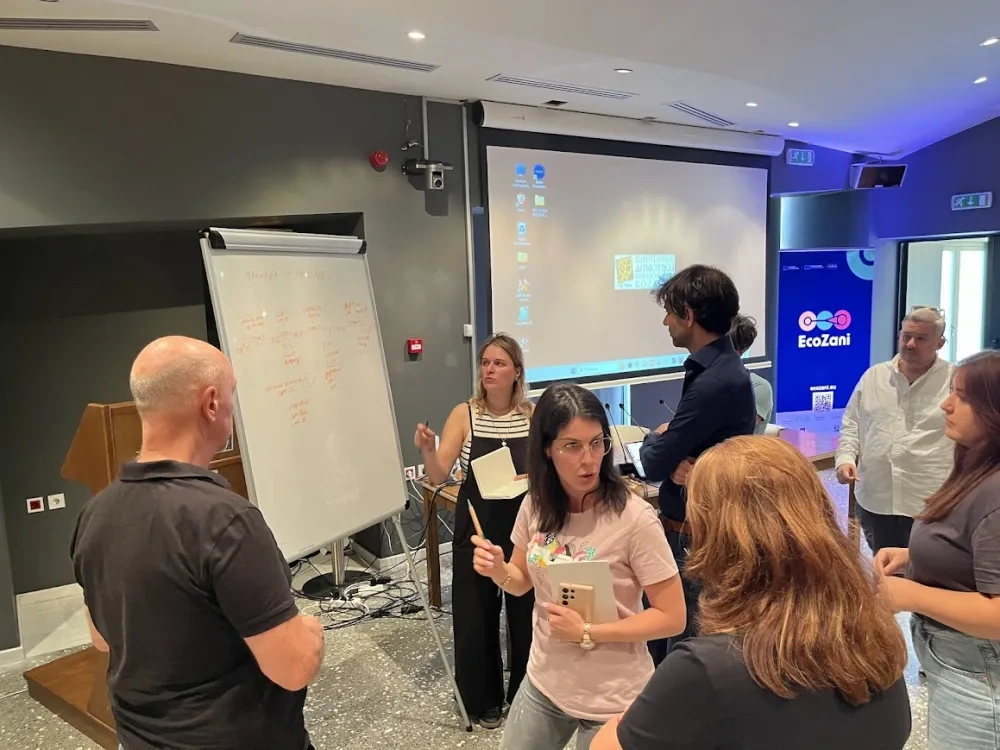 Participants engage in an animated group discussion during an EcoZani workshop, standing around a flipchart and exchanging ideas in an interactive working session in a conference room.